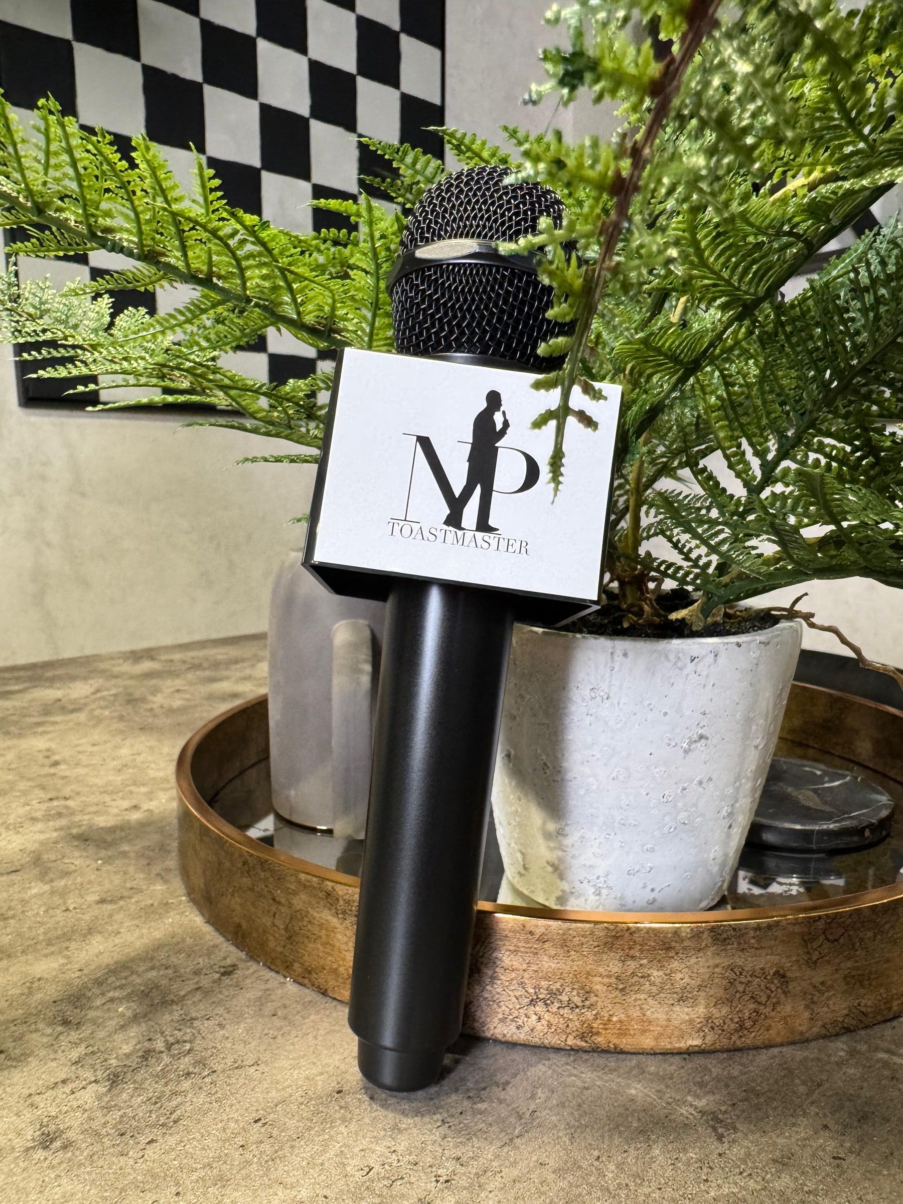 Niral's black microphone with an NP Toastmaster branded mic flag, resting on a gold tray beside a fern