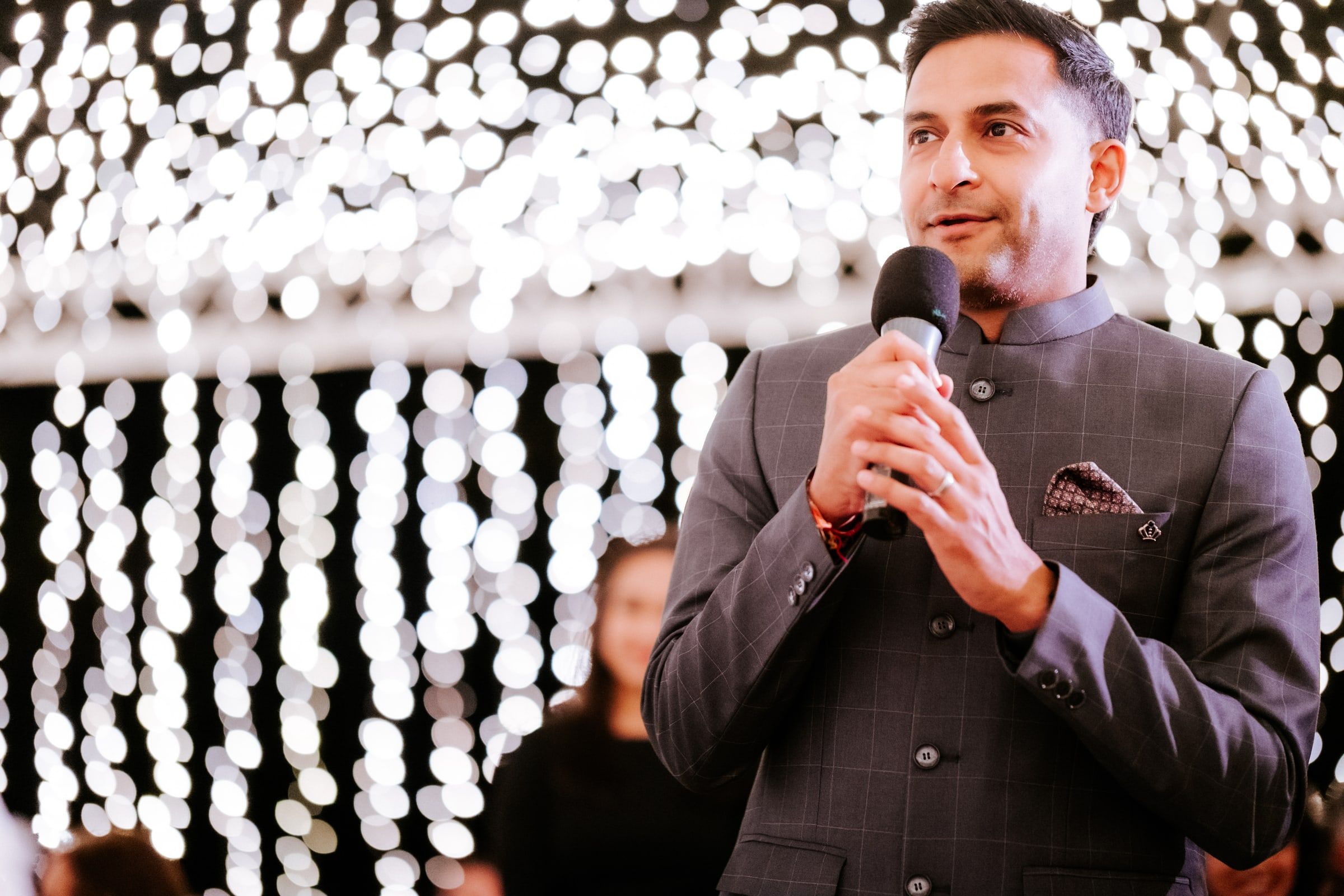 Niral in a grey bandhgala speaking into a microphone in front of a curtain of fairy lights at an evening reception
