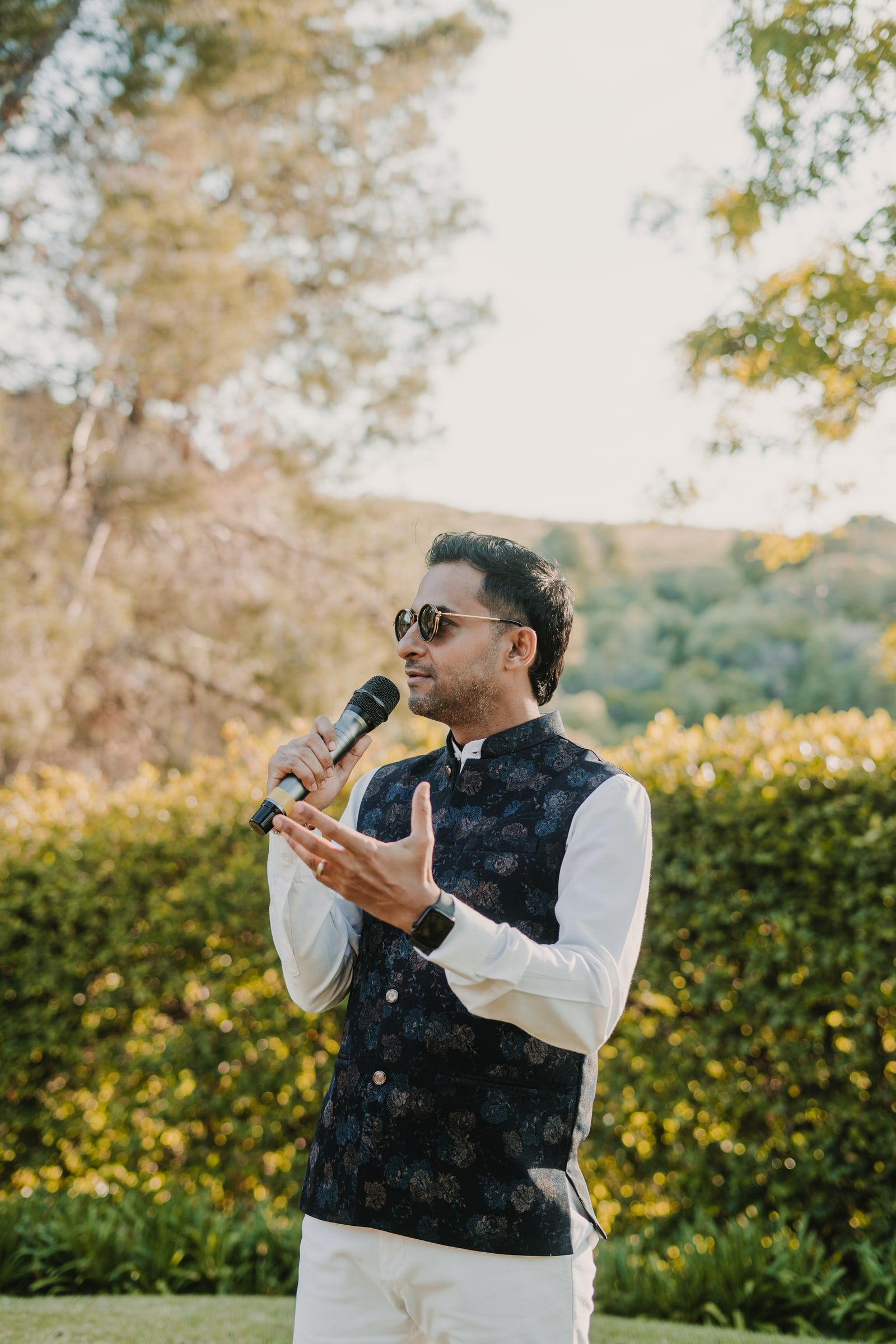 Niral in a navy Nehru jacket and sunglasses, mic in hand, gesturing to guests at a sunny garden wedding