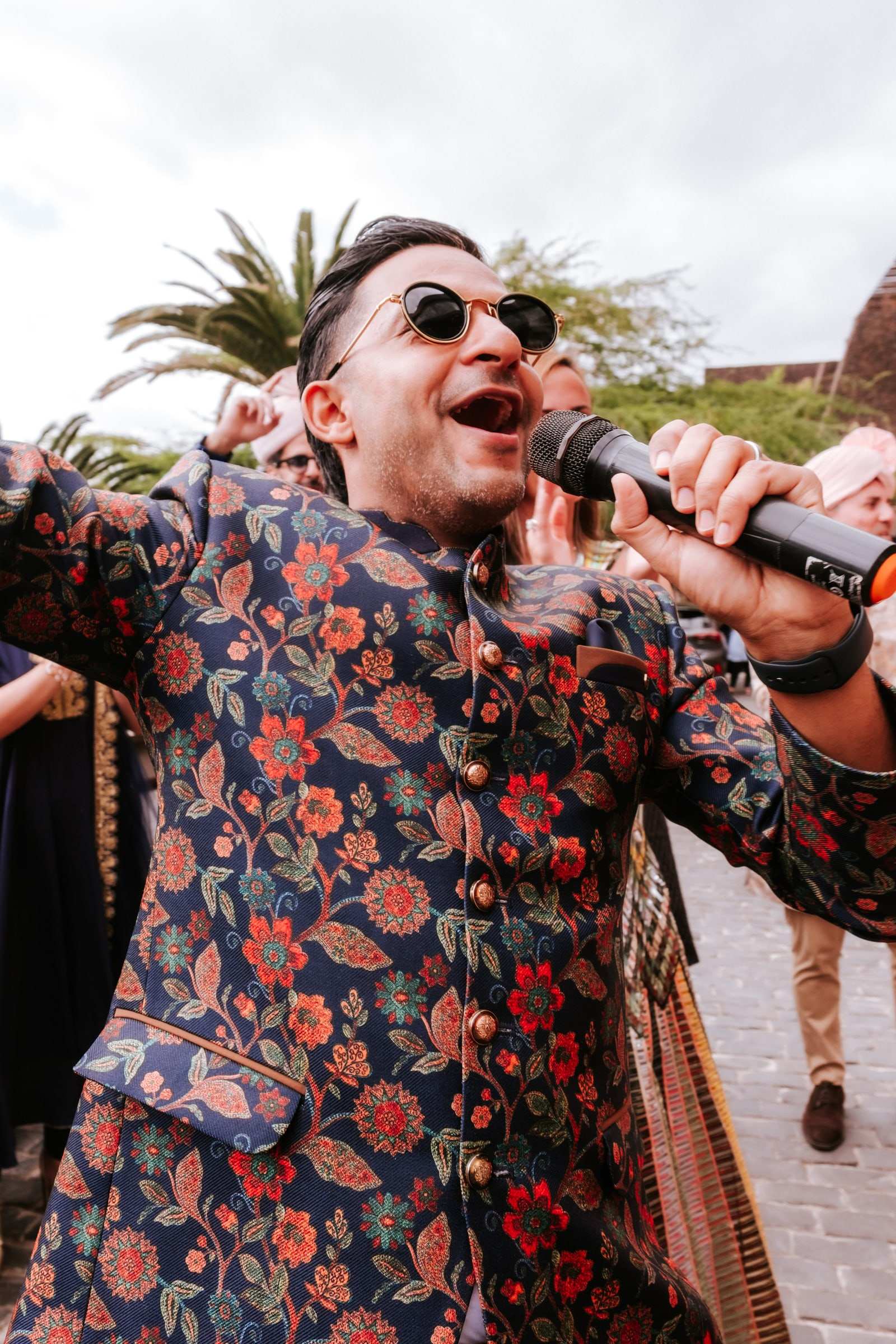 Niral in a floral sherwani and sunglasses leading a baraat procession, singing into the microphone