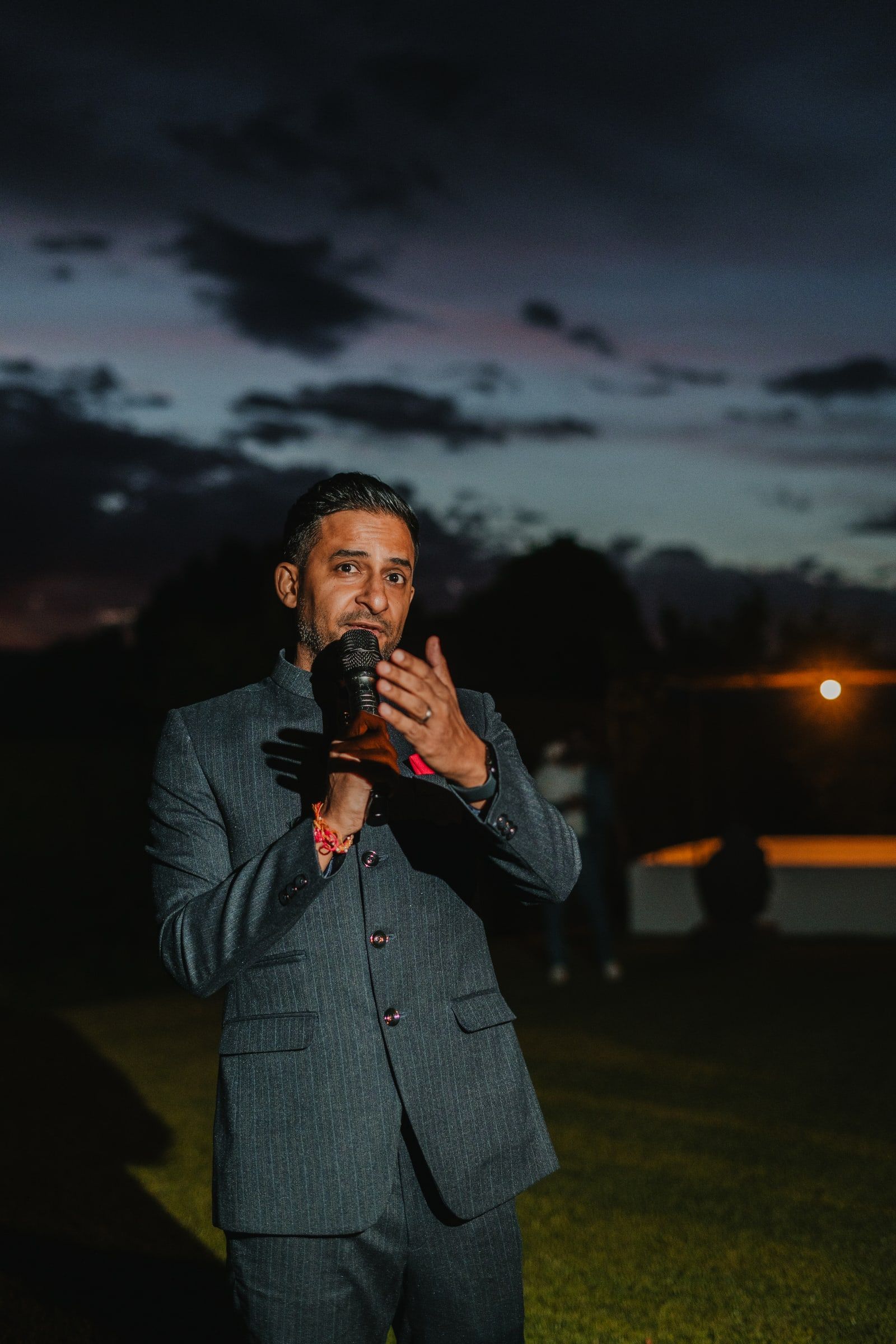 Niral in a charcoal suit speaking into a microphone under a dusky evening sky