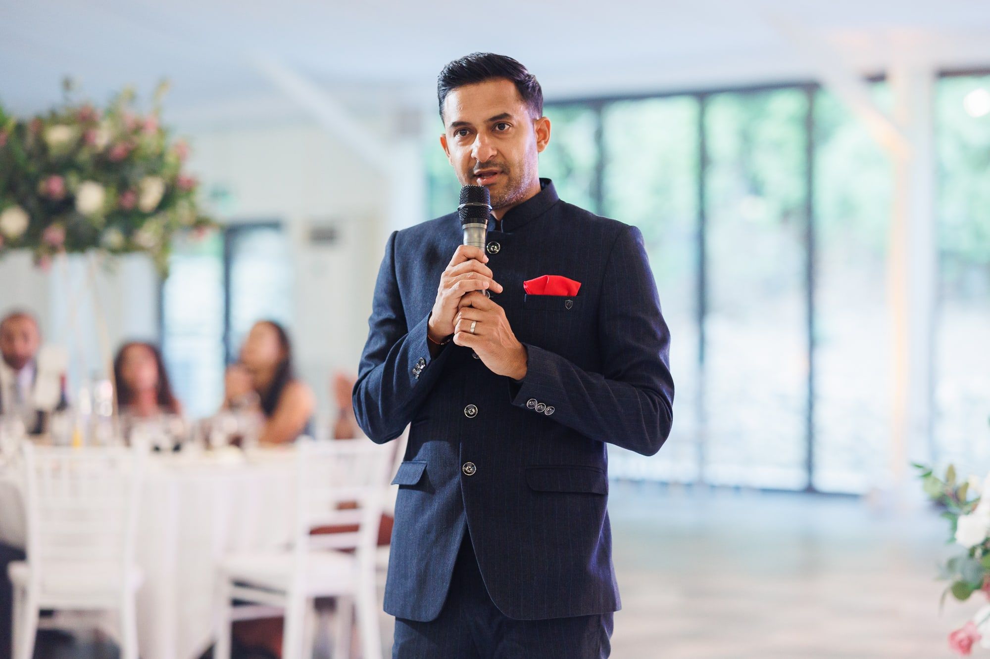 Niral in a navy bandhgala with a red pocket square addressing guests seated at a wedding breakfast